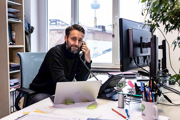 Mobilitätsplaner arbeitet im Büro am Schreibtisch, telefoniert und blickt auf mehrere Monitore.