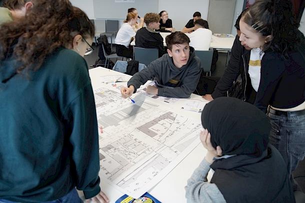 Ein Tisch mit Studierenden, die mit Hilfe von verschiedenen Planungsskizzen die Stadt theoretisch planen.