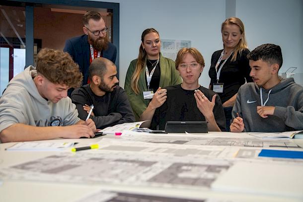Studierende und Fachleute mit AGFS-Namensschildern diskutieren an einem Tisch mit großformatigen Planzeichnungen