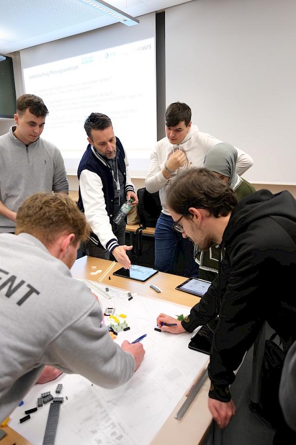 Eine Gruppe von sechs Personen arbeitet an einem Projektplan auf einem Tisch ausgebreitet mit Lego-Bausteinen und bespricht die aktuellen Ergebnisse.